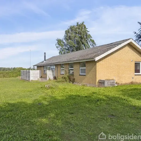 En enkeltstående gul murstensbungalow står på en stor, grøn græsplæne. Et hegn løber langs venstre side, og himlen er klar og blå med enkelte skyer.