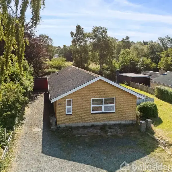Et gult murstenshus står i centrum, omgivet af en grusvej og grønne træer. Huset har et skråt tag og store vinduer. Området er åbent og grønt.