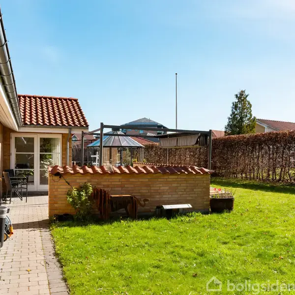 Hus i rød mursten med terrasse, hvor havemøbler står op af muren; græsplæne omgiver området, og hække markerer haven i et solrigt boligområde.