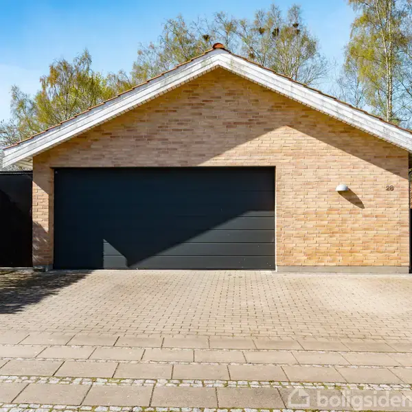 Garage med brun murstensfacade og sort port står på en flisebelagt indkørsel omkranset af hæk. Nummer 10 ses på facaden. Baggrunden er skovklædt.