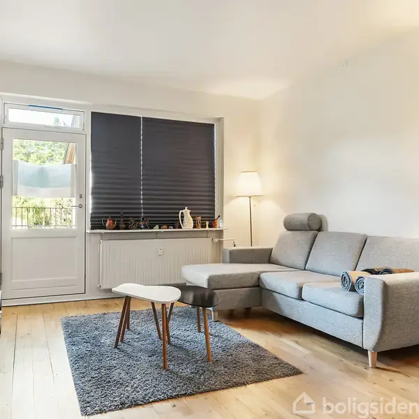 Sofa står i en lys stue med trægulv, akkompagneret af to lænestole og et sofabord på et tæppe. En lampe lyser hjørnet, og et vindue med persienner er i baggrunden.