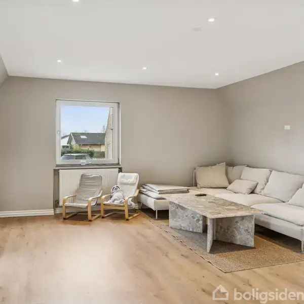 Lys stue med en beige hjørnesofa og sofabord. Væghængt fladskærms-TV til venstre. To stole ved vindue med udsigt til et hus. Trægulv og lyse vægge.