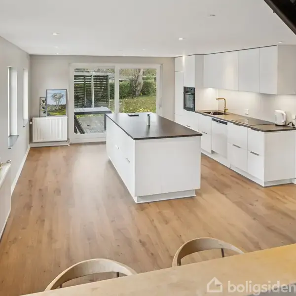 Køkken med hvidt inventar og mørk bordplade, centreret køkkenø. Store vinduer slipper lys ind, og trægulv breder sig gennem rummet. Moderne og rummeligt design med udsigt til have.