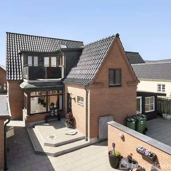 En rød murstensvilla med sort tag beliggende i et beboelsesområde. Huset har en stor terrasse med krukker og en have afgrænset af et stakit. Solskinsvejr opløfter scenen.
