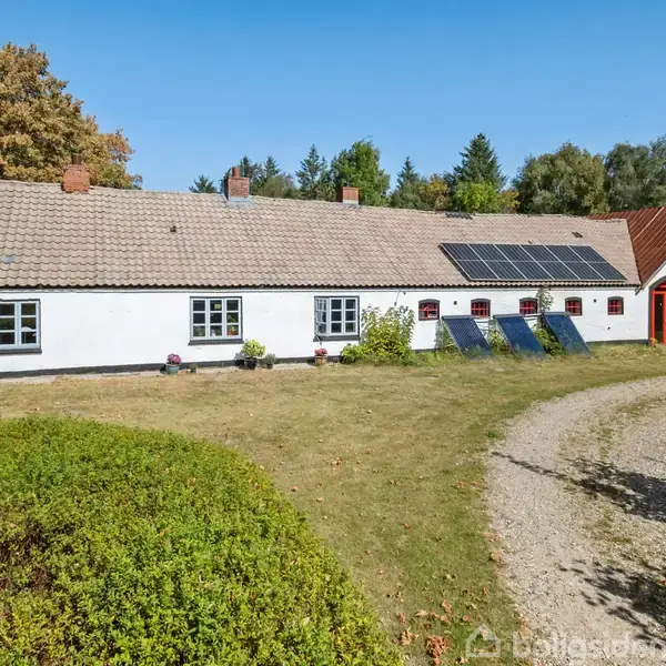 Et hvidt, langstrakt hus med rødt tag og solpaneler står i en grøn have med grussti. Omkranset af træer i efterårsfarver i et landligt område.