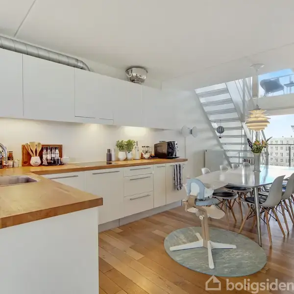 Et moderne køkken med hvidt interiør og træbordplader. Spisebord med stole foran en glasvæg ved siden af en trappe. Lys træparketgulv og ovenlys giver rummet et lyst udseende.
