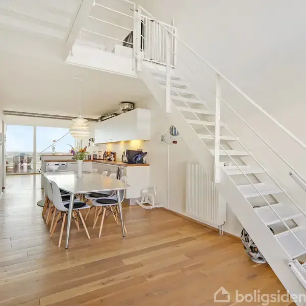 Moderne, lyst køkken-alrum med hvidt bord og seks stole. Trætrappe fører til øverste etage. Store vinduer giver udsigt og lysindfald. Åbne hylder og planter skaber en hyggelig atmosfære.