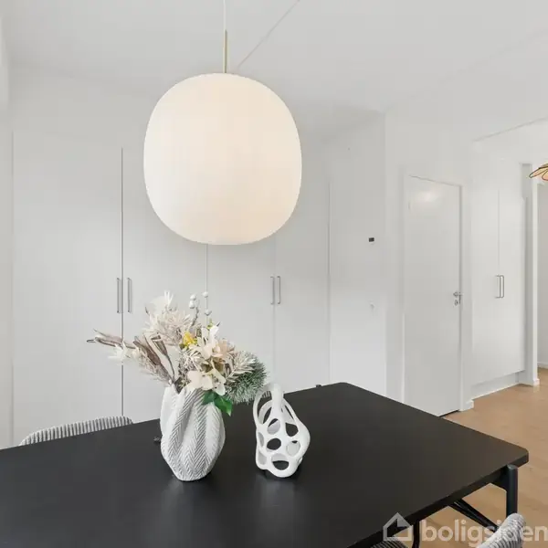 En sort bordplade med to hvide vaser (den ene med blomster) står under en stor hvid lampe. Baggrunden er et lyst rum med skabe og en åben dør.