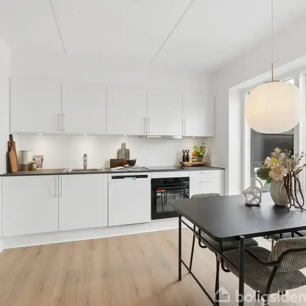 Et moderne køkken med hvide skabe udfører funktioner som opbevaring og madlavning. Et elegant spisebord er omgivet af stole, mens en stor lampe hænger over det. Der er blomster i en vase på bordet.