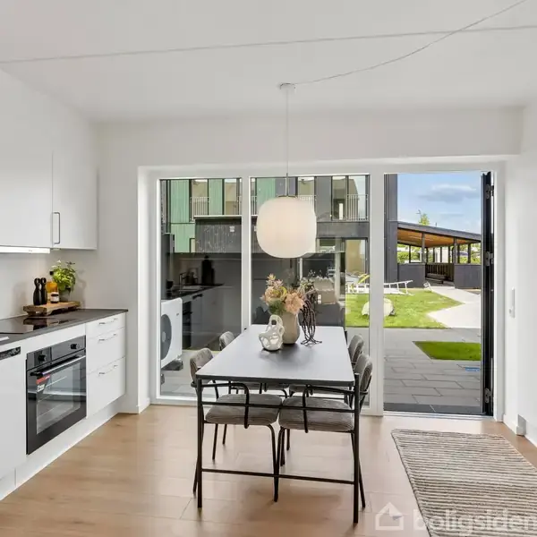 Et moderne køkken med hvidt interiør og træbord er centralt placeret. Bordet har en vase med blomster, og store vinduer åbner ud til en solrig terrasse og have.
