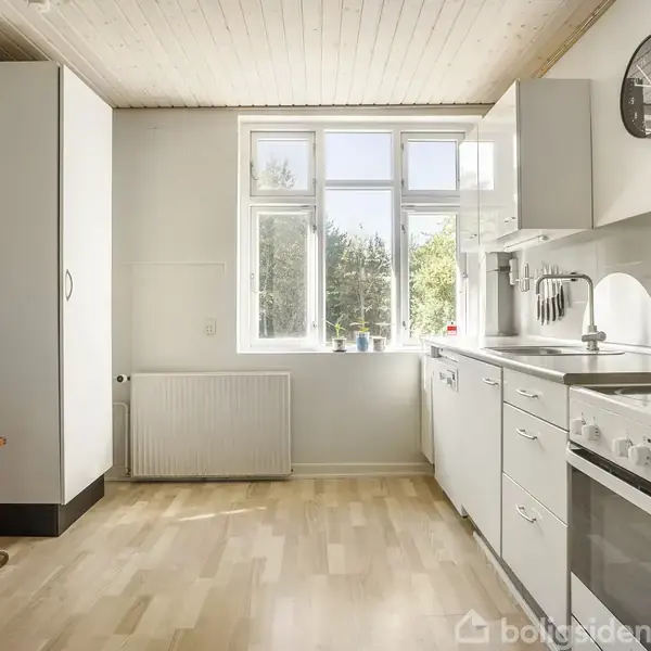 Køkken borde med hvide skabe står ved højre side. Ovn og vask er integreret. Et træbord står ved venstre side. Et vindue viser træer udenfor. Ingen tekst synlig.