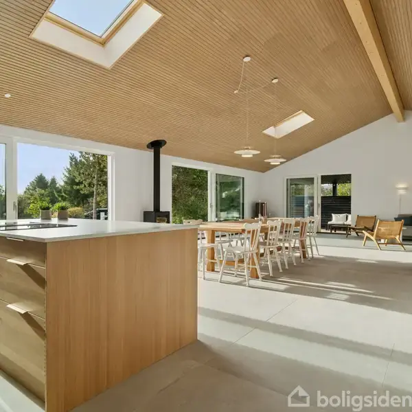 Et moderne køkkenalrum med et træloft og store vinduer. Køkkenøen står midt i rummet, omgivet af spisebord og stole. Grønne planter tilføjer en naturskøn atmosfære.