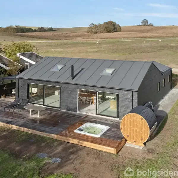 Et moderne, sort træhus med store vinduer står på en træterrasse. En sauna i tøndeform står ved siden af en jacuzzi. Omgivelserne er åbne marker og landskab.