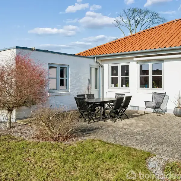 Terrassebord med stole står på en stenterrasse foran et hvidt hus med rødt tegltag. Til venstre er en busk, og der er en grill til højre.