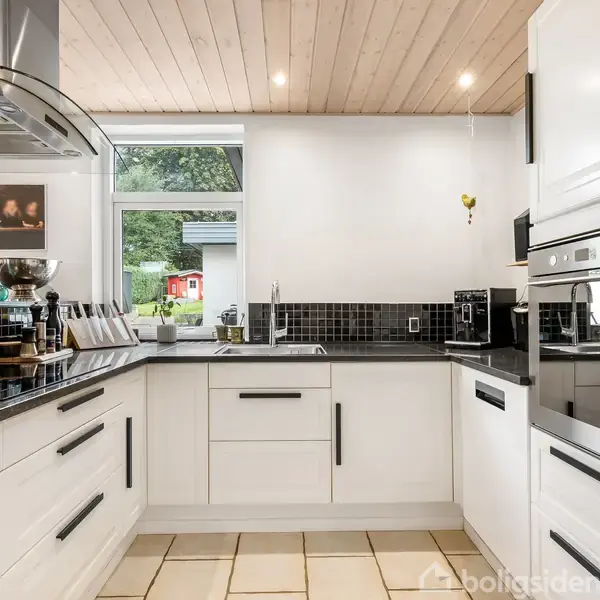 Et moderne køkken med hvide skabe og sorte bordplader. Indbyggede apparater er til stede, og der er et stort vindue, der giver udsigt til en have med græs og et lille rødt hus.