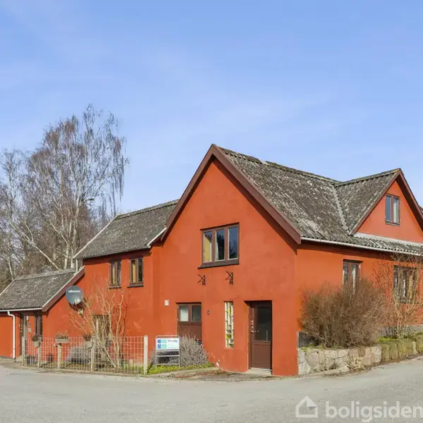 Rødt hus står stille ved vejkryds; omgivet af asfaltvej, træer og nabohus med stråtag. Tydelige vinduer og dør på facaden, placeret i landlig, åben omgivelser med blå himmel.