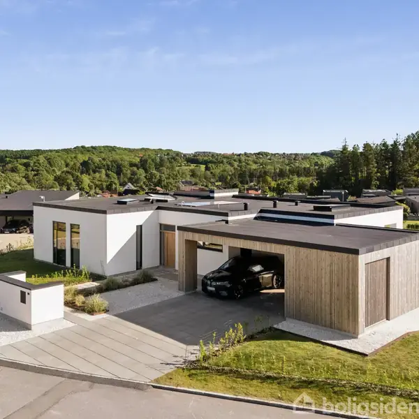 Moderne hus med fladt tag og carport, der beskytter en sort bil. Omgivet af grønne områder og ligger i et boligkvarter med flere huse på en solrig dag.