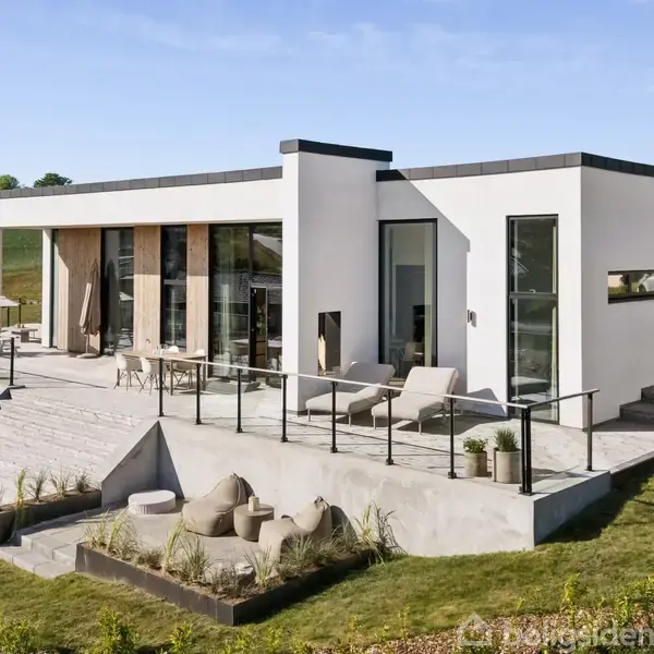 Moderne hvid villa med fladt tag står på en forhøjet terrasse omgivet af glasrækværk. Ligger i en grøn have ved siden af store græsarealer under en blå himmel.
