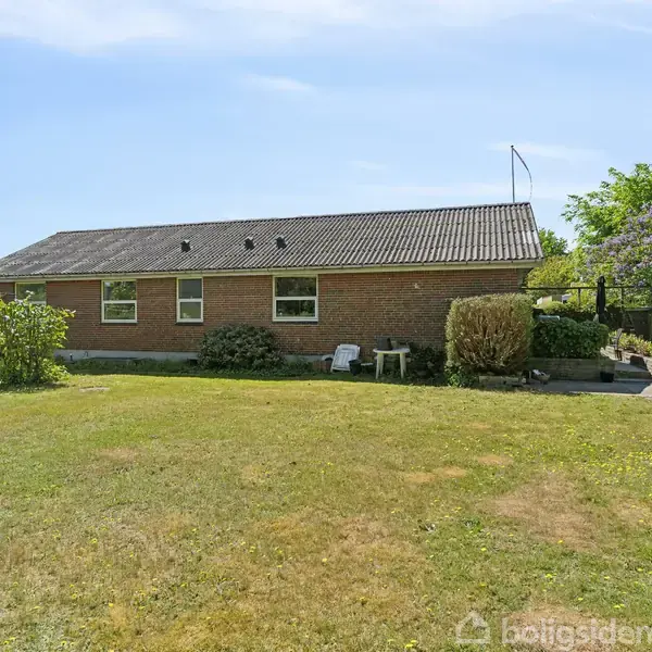 En lav murstensbygning står stille. Foran er en græsplæne omkranset af buske og havemøbler, der giver et hyggeligt indtryk. Himlen er blå og klar.