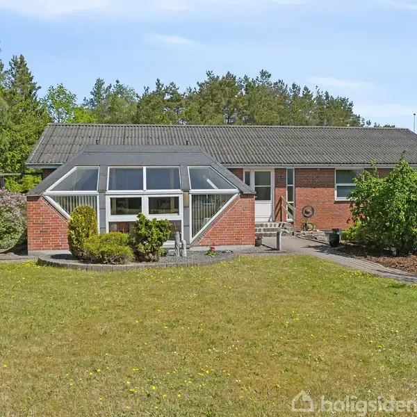 Et enfamilieshus med røde mursten står stille i en velplejet have omkranset af buske. Foran huset er en stor græsplæne, og i baggrunden ses en række træer.