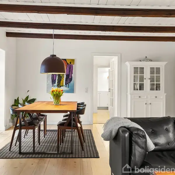 En spisebordsgruppe med træbord og sorte stole står på et tæppe. Over bordet hænger en lampe. Rummet har hvide vægge, bjælkelofter og en sort lædersofa i forgrunden.