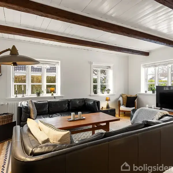 En stue med sorte lædersofaer omkring et træbord. Rummet har hvidmalede vægge, store vinduer, og synlige træbjælker i loftet. En gulvlampe står ved siden af sofaen.