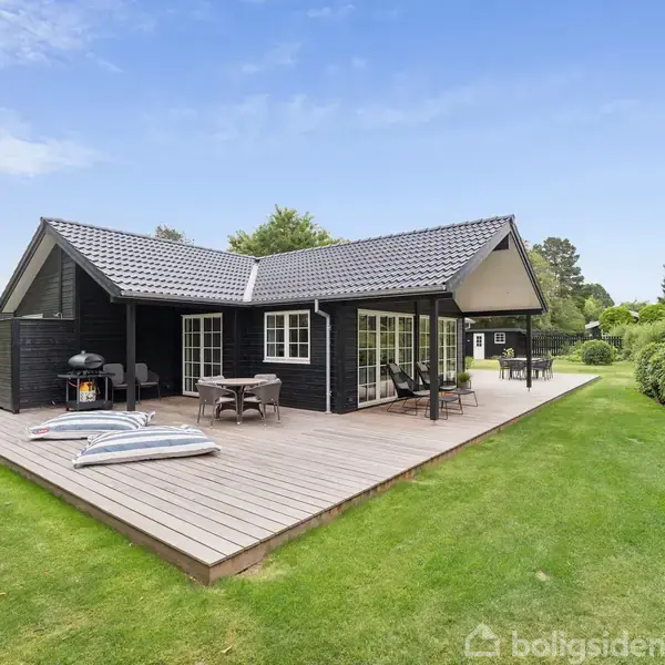 Sort træhus med terrasse, der har to solsenge og et bord med stole. Huset ligger i en frodig have med grønne træer og buske.