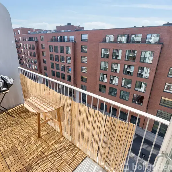 Balkonen har et lille rundt bord og en grill, der er dækket med et sort betræk. Omgivet af høje røde murstensbygninger og blå himmel.
