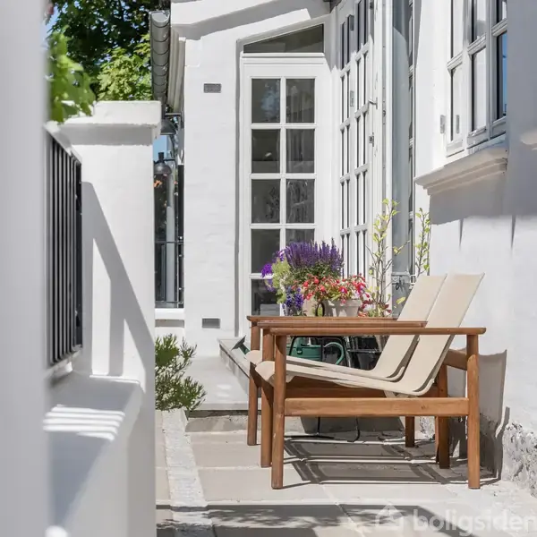 En træstol og et bord står på en solrig terrasse foran et hvidt murstenshus med store vinduer. Blomsterdekorationer tilfører farve til omgivelserne.