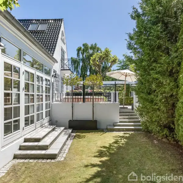 Hus med store vinduer åbner ud til en lille terrasse og have. Trapper fører til en hævet terrasse med parasol. Haven er omgivet af høje træer.