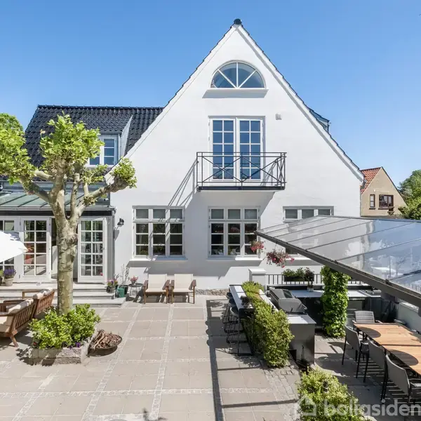 Hvidt hus med spidst tag, omgivet af stor terrasse. Terrassebord og stole placeret under åben himmel. Grønne planter og solbeskinnet, blå himmel i baggrunden.