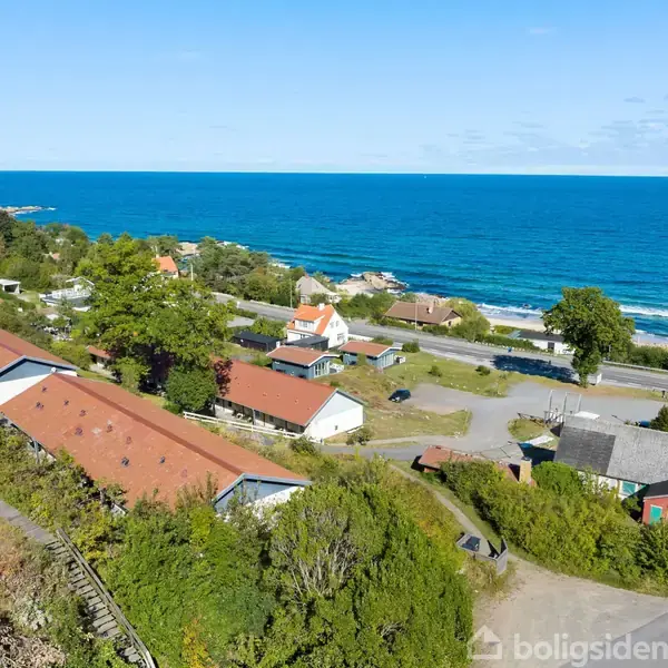 Række af huse med røde tage ligger stille, omgivet af grøn vegetation, tæt på en bred vej ved kysten med udsigt over det blå hav under en skyfri himmel.