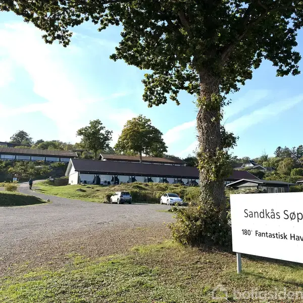 Bygninger stående ved siden af en grusvej omkranset af træer; en skilt med teksten "Sandkås Søpark, 180° Fantastisk Havudsigt" er placeret i forgrunden.