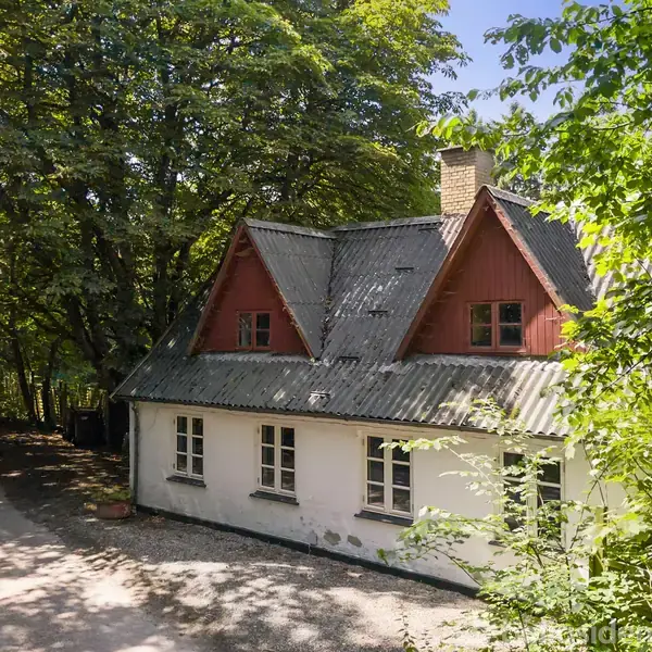 Et charmerende hvidt hus med røde gavle og skifertag står fredeligt omgivet af frodige træer. En grusvej løber lige ved siden af huset.