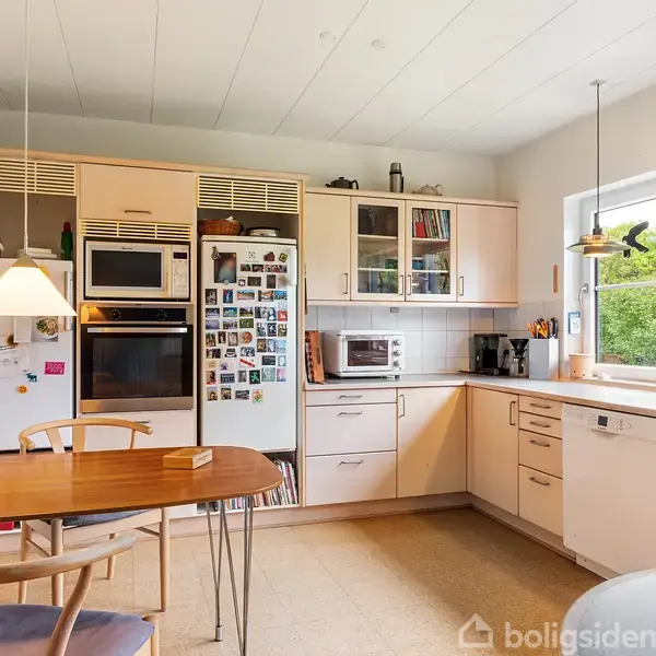 Køkken med træskabe og hvidevarer; en ovn, mikroovn og køleskab med magneter. Et lille bord med stole, lamper over bordet. Vindue med udsigt til have, vægur ved siden.