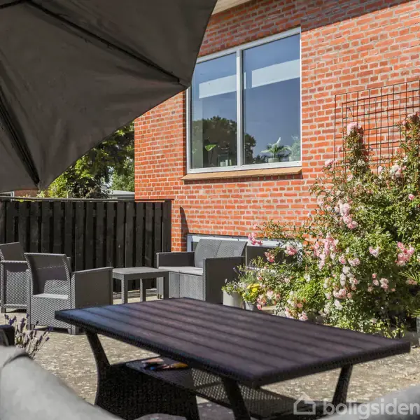 En terrasse med sorte havemøbler arrangeret tæt på et rødt murstenshus. Pink blomster vokser op ad et espalier ved siden af huset, mens en stor parasol skygger området.