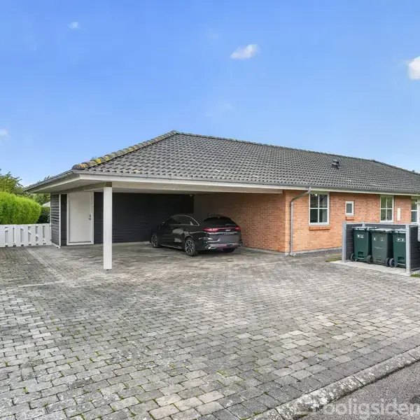 Et murstenshus med fladt tag står stille. En bil er parkeret under en carport til venstre. Området omkring er en brolagt indkørsel med grønne hække som baggrund.