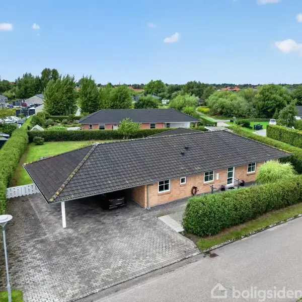 Et gult murstenshus med et lyst tegltag har en carport ved siden af sig. Huset er omgivet af en velholdt have med grønne hække i et boligområde.