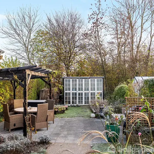 En have med et sæt havemøbler under en pergola, omgivet af forskellige planter. Et drivhus ses i baggrunden, der grænser op til en skovklædt baggrund.