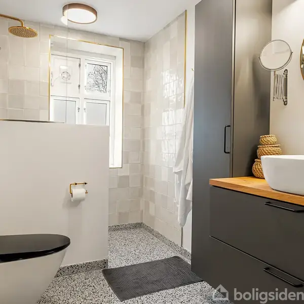 Toilet med sorte sæde, placeret ved væg med papirholder, i moderne badeværelse med brusekabine, gråskabsarrangement, rundt spejl og vaskeskål, i lyst rum med flisegulv.