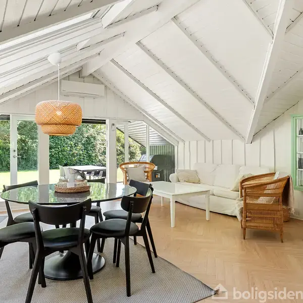 Stue med spisebord og stole i forgrunden, sofa og stol ved siden af. Store vinduer og dør åbner mod grøn have. Loftet er hvidmalet med synlige bjælker.
