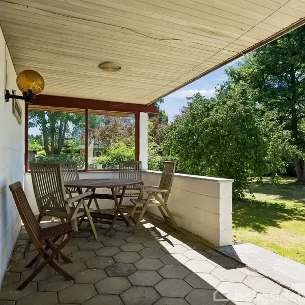 En overdækket terrasse med træmøbler og seks klapstole omkring et bord, beliggende ved siden af en glasdør. I baggrunden ses en grøn have med træer og buske.