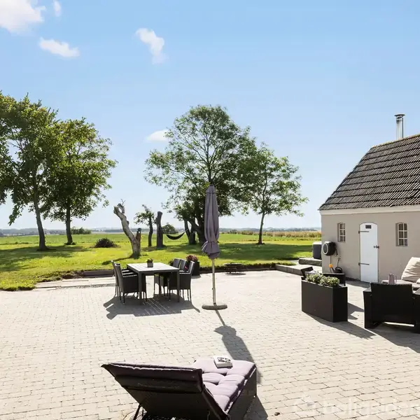 Terrasse med flisebelægning og havemøbler står mellem to hvide bygninger. I baggrunden ses græsplæne med enkelte træer, der giver udsigt til marker og en klar blå himmel.
