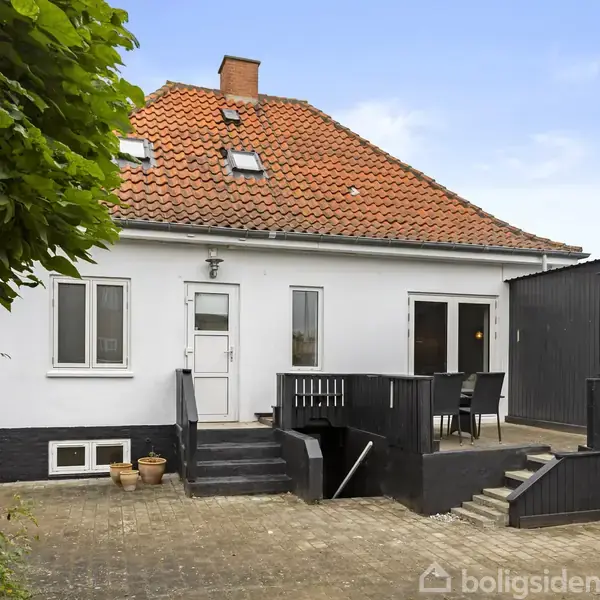 Hvidt hus med rødt tag står stille i en gård med flisebelægning. Terrassen har sorte møbler, omkranset af grønne planter og hegn.