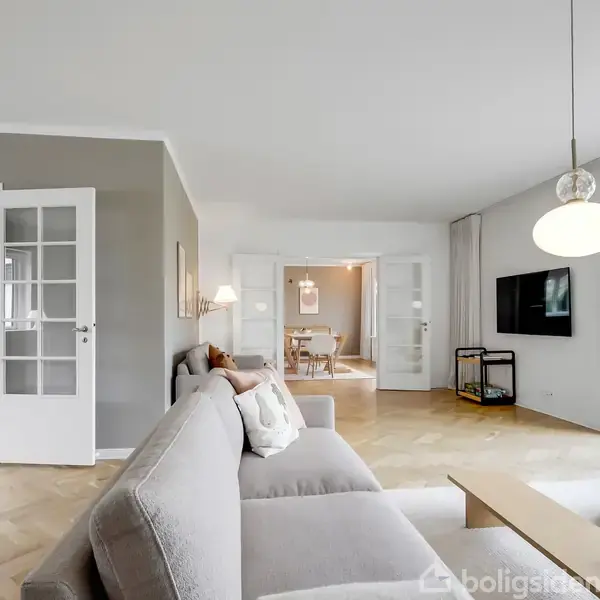 Grå sofa står i en lys stue med parketgulv, mod en åben dør til en spisestue. Fladskærms-TV hænger på væggen. Store vinduer giver udsigt til grønne træer.