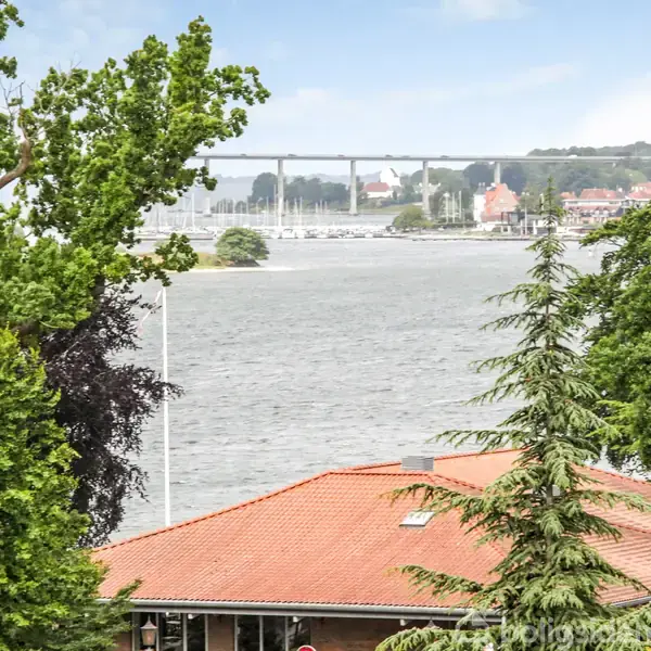 En rødtagget bygning står mellem frodige træer ved en flod. Bagved ses en lang bro og byens skyline i det fjerne under en klar himmel.