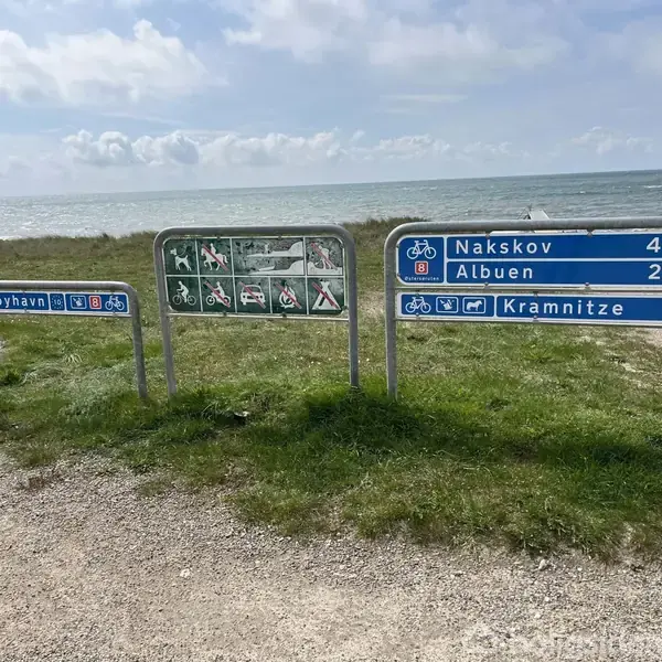 Tre vejskilte står på en grusvej ved havet. Teksten angiver afstande: Nakskov 44, Albuen 25, Kramnitze 5, Rødbyhavn. Ikoner indikerer cykelruter og faciliteter.