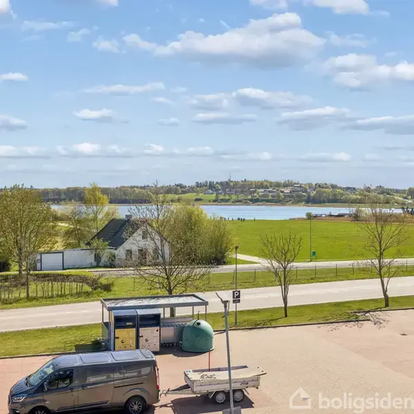 En grå varevogn med trailer holder parkeret på en asfaltplads foran en busstoppested. I baggrunden ses en grøn mark, træer og en sø under en blå himmel med skyer.