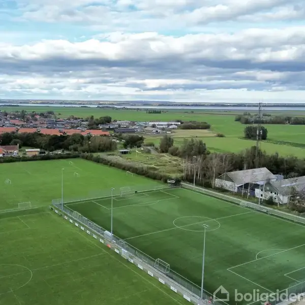 Fodboldbane ligger stille mellem en lille landsby og grønne marker, med huse i baggrunden og en himmel dækket af skyer. Ingen tekst fundet i billedet.