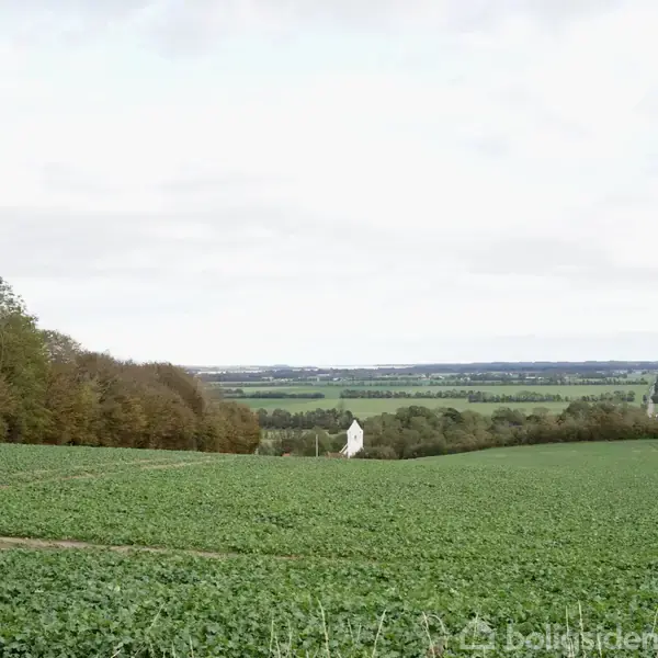 En hvid bygning står i en grøn mark. Den er omgivet af landskab med skove og åbne marker under en lys himmel.